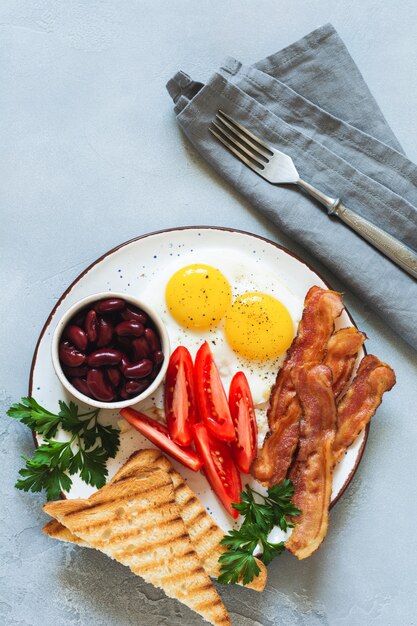 Traditionelles englisches Frühstück mit Spiegeleiern, Würstchen, Bohnen, gegrillten Tomaten und Speck auf grauem Betonhintergrund. Ansicht von oben