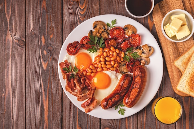 Traditionelles englisches Frühstück mit Spiegeleiern, Würstchen, Bohnen, Pilzen, gegrillten Tomaten und Speck auf Holzhintergrund. Ansicht von oben