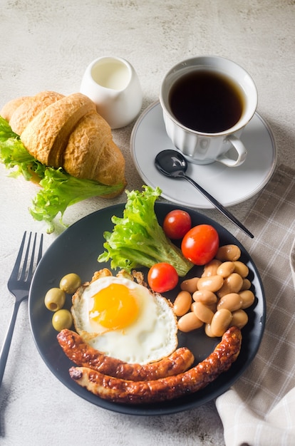 Traditionelles englisches Frühstück mit Spiegeleiern, Würstchen, Bohnen, Tomaten auf einem Teller, Croissant und Kaffee auf grauem Hintergrund. Von oben betrachten