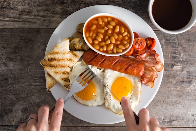 Traditionelles englisches Frühstück mit Spiegeleiern, Würsten, Bohnen, Pilzen, gegrillten Tomaten und Speck auf Holzoberfläche