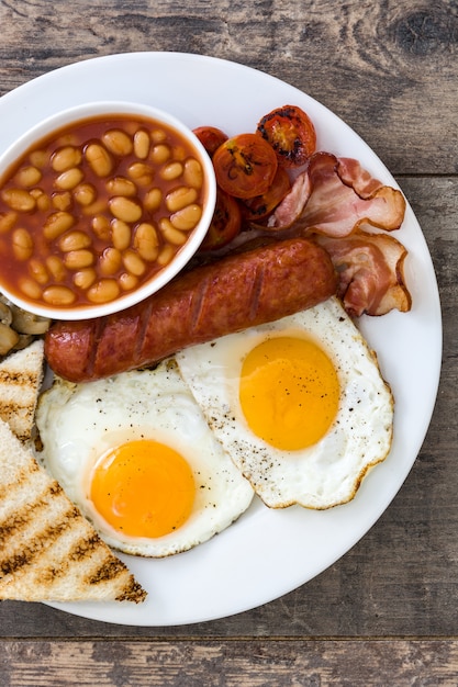 Traditionelles englisches Frühstück mit Spiegeleiern, Würsten, Bohnen, Pilzen, gegrillten Tomaten und Speck auf Holzoberfläche