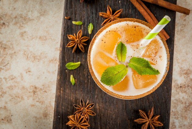 Traditionelles indisches Getränk ist Eistee oder Chai Masala mit Eiswürfeln aus Chai, Milch und Minze. Mit gestreiften Strohhalmen auf einem Holzbrett. Auf hellbeigem Steintisch. Copyspace Draufsicht