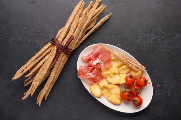 Traditionelles italienisches Essen ist Grissini-Brot mit Schinken, Käse und Tomaten mit Kräutern auf einem Teller auf dunklem Hintergrund.