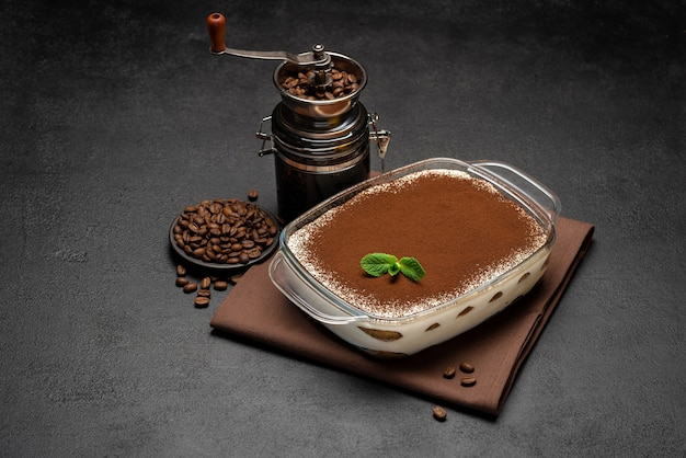 Traditionelles italienisches Tiramisu-Dessert in Glasbackform und Kaffeemühlenmühle auf Betonhintergrund oder -tisch