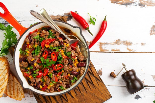 Traditionelles mexikanisches Essen - Chili con Carne mit Hackfleisch und Gemüseeintopf in Tomatensauce in einer gusseisernen Pfanne auf hellgrauem Schiefer oder Betonhintergrund. Draufsicht mit Kopienraum