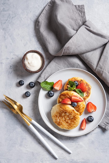 Traditionelles russisches Gericht zum Frühstück Syrniki Hüttenkäse-Pfannkuchen mit Sauerrahm und frischen Beeren auf blauem Hintergrund mit Geschirr und Serviette Draufsichtbild Vertikale Ausrichtung