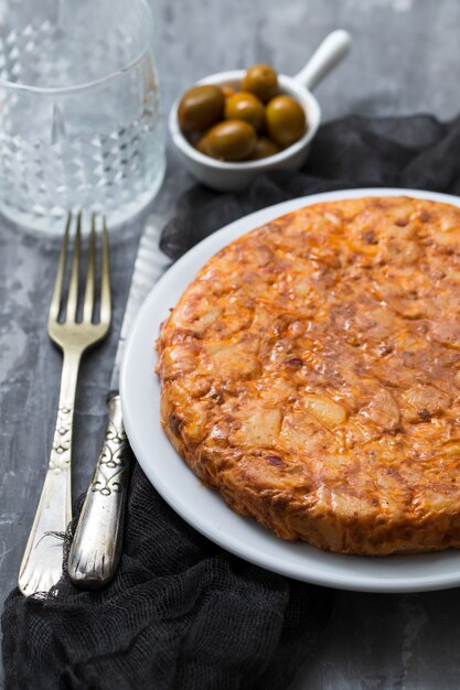 Traditionelles spanisches Essen Tortilla mit Kartoffel auf weißem Teller und Oliven in der kleinen Schüssel auf keramischem Hintergrund.