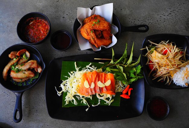 Foto traditionelles thailändisches essen mit lachs, scharfem gebratenen hühnerfleisch, schweinefleisch, papaya-salat auf schwarzem teller auf dem tisch