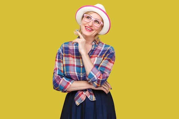 Träume von etwas Schönem. Porträt einer modernen, stilvollen, reifen Frau mit Zahnsmiley im lässigen Stil mit Hut, der steht, wegschaut und träumt. Indoor-Studioaufnahme isoliert auf gelbem Hintergrund