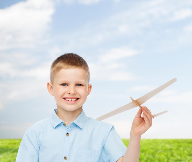 Träume, Zukunft, Hobby, Natur und Kindheitskonzept - lächelnder kleiner Junge, der Holzflugzeugmodell in der Hand über natürlichem Hintergrund hält