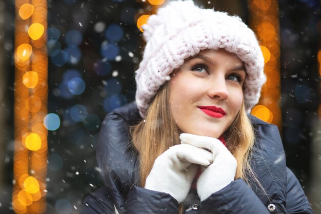 Träumende blonde Frau trägt Strickmütze, die bei Schneefall auf der Wintermesse spazieren geht. Leerer Platz für Text