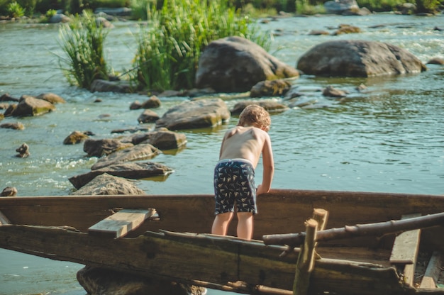 Träumender Junge im alten Boot an der Seeküste. Netter kleiner Junge, der im alten Holzboot auf der Natur sitzt.