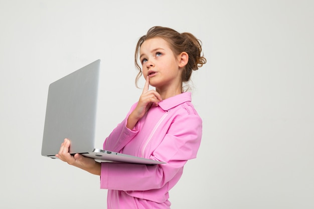 Träumendes Mädchen in einem rosa Anzug hält einen Laptop in seinen Händen an einer weißen Wand