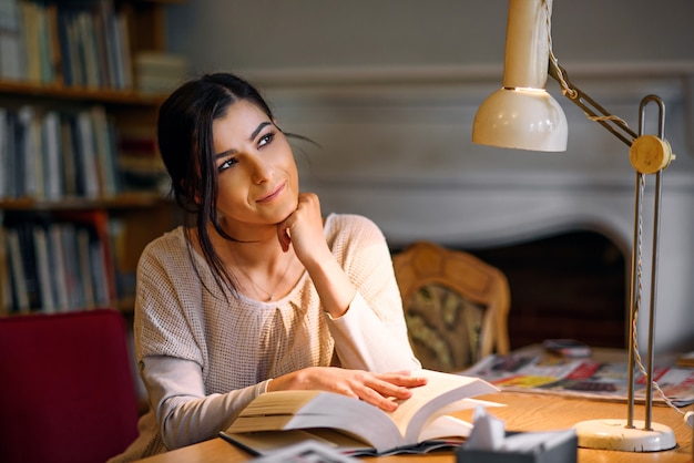 Träumerisches hübsches und enthusiastisches Studentenmädchen-Lesebuch in der Universitätsbibliothek unter einer Tischlampe