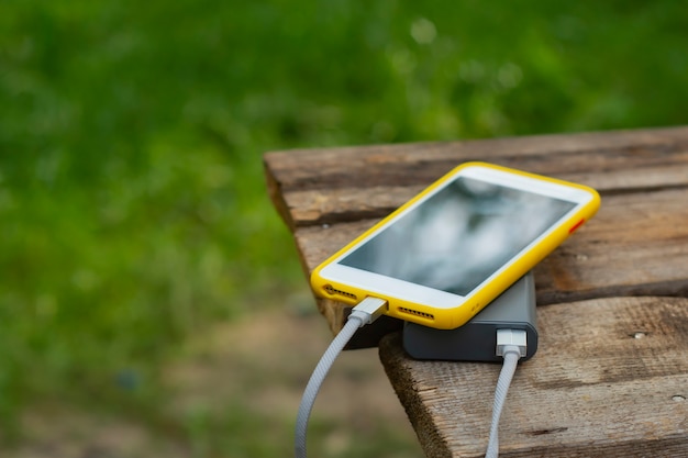 Tragbares Reiseladegerät. Power Bank lädt ein Smartphone auf einem Holztisch im Hintergrund der Natur auf. Konzept zum Thema Tourismus.