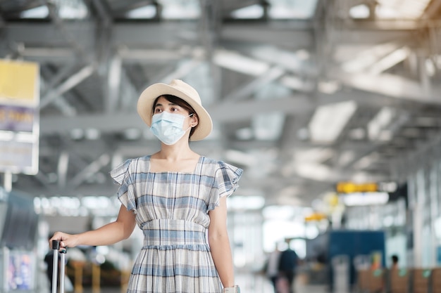 Tragende Gesichtsmaske der jungen Frau mit Gepäck, das Flugzeit im Flughafen prüft