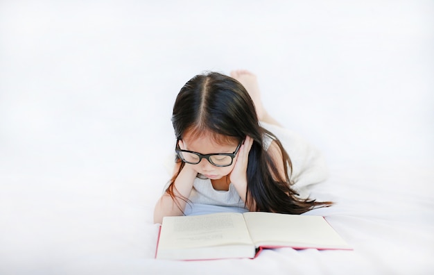 Tragende Gläser des kleinen asiatischen Kindermädchens, die das gebundene Buch liegen auf Bett gegen weißen Hintergrund lesen.