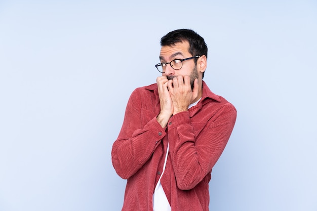 Tragende Kordjacke des jungen kaukasischen Mannes über der blauen Wand nervös und erschrocken, Hände zum Mund setzend