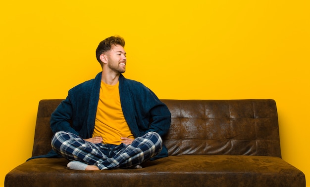 Tragende Pyjamas des jungen Mannes, die glücklich, nett und überzeugt schauen, stolz lächeln und schauen, um mit beiden Händen auf Hüften mit Seiten zu versehen. auf einem Sofa sitzen