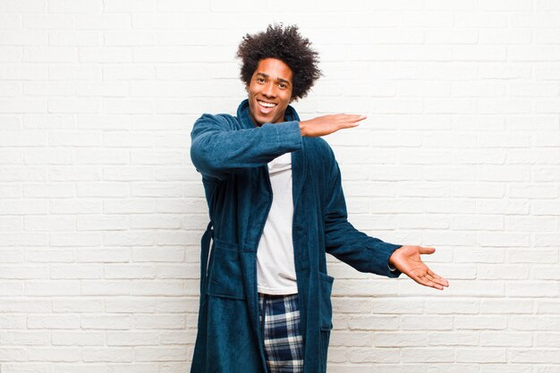 Tragende Pyjamas des jungen schwarzen Mannes mit Kleid lächelnd, glücklich, positiv und zufrieden, Gegenstand oder Konzept auf Kopienraum gegen Backsteinmauer halten oder zeigend