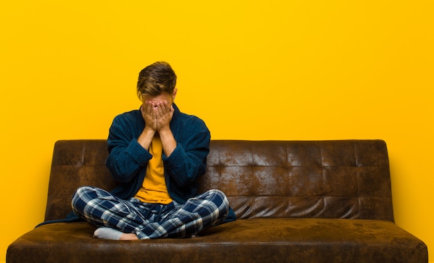 Tragender Pyjama des jungen Mannes, der traurig, frustriert, nervös und deprimiert sich fühlt und das Gesicht mit beiden Händen bedeckt und schreit. auf einem Sofa sitzen