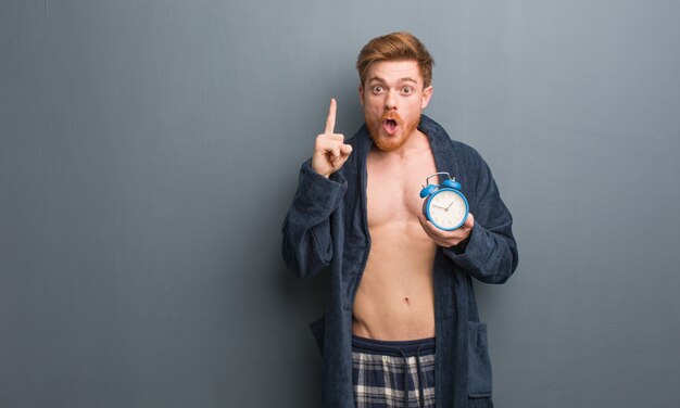 Tragender Pyjama des jungen Rothaarigemannes, der eine großartige Idee, Konzept der Kreativität hat. Er hält einen Wecker.