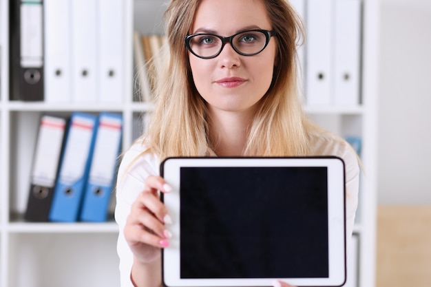 Tragendes Glasporträt der schönen Geschäftsfrau im Büro, das in der Hand eine Tablette sitzt am Tisch lächelt und betrachtet den Kameralehrer hält, drückt den Erfolg aus, der die Testpapiere überprüft.