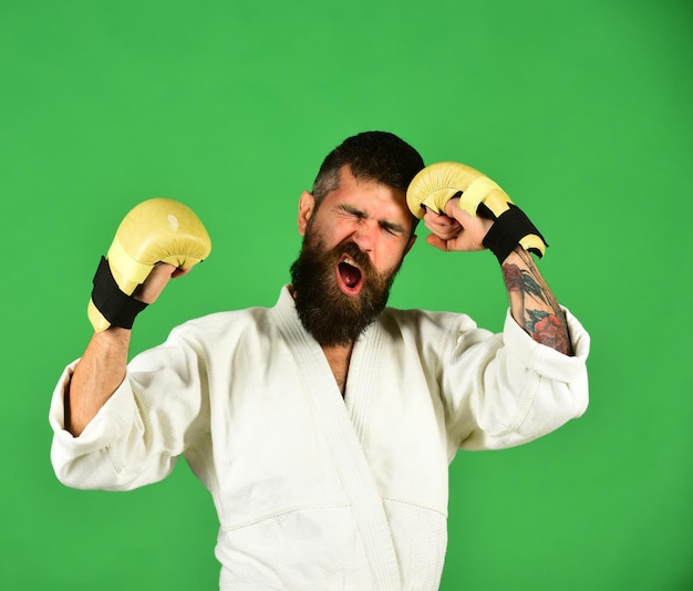 Trainings- und Kampfkonzept. Kampfmeister schlägt sich mit der Faust. Karate-Mann mit leidendem Gesicht in Uniform und goldenen Boxhandschuhen. Mann mit Bart im weißen Kimono auf grünem Hintergrund.