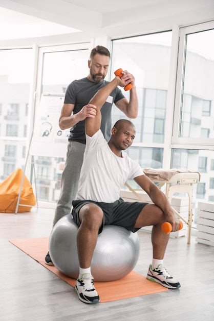 Trainingsgeräte. Netter junger Mann, der auf dem Medball sitzt und Hanteln hebt