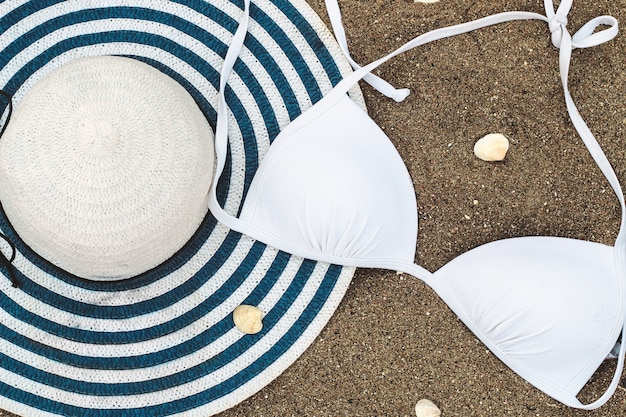 El traje de baño de mujer blanco se encuentra en la arena con conchas marinas junto con un sombrero para el sol.