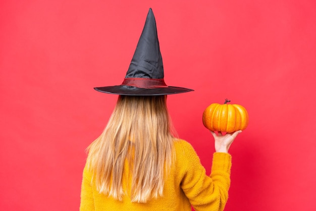 Traje de mujer caucásica joven como bruja sosteniendo una calabaza aislada sobre fondo rojo en posición trasera