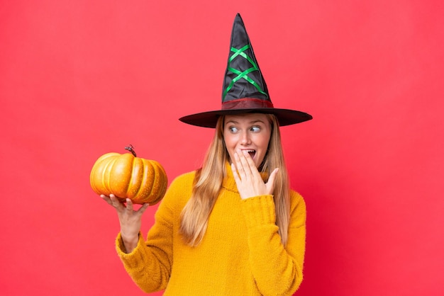 Traje de mujer caucásica joven como bruja sosteniendo una calabaza aislada sobre fondo rojo con sorpresa y expresión facial conmocionada