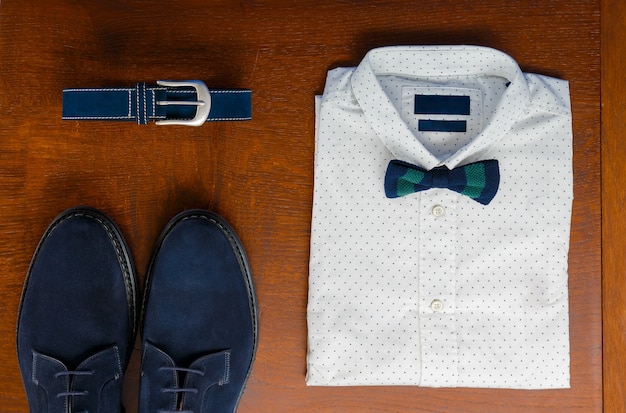 Trajes masculinos, camisa de bolinhas brancas com gravata borboleta, cinto azul e sapatos em fundo marrom. Acessório de casamento dos homens.