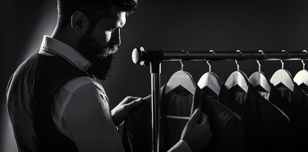 Trajes masculinos colgados en fila. Ropa de hombre, boutiques. Sastre, sastrería. Traje de hombre con estilo. Traje de hombre, sastre en su taller. Hombre guapo con barba en traje clásico. En blanco y negro.