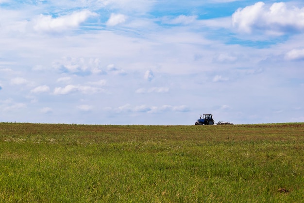 Traktor, der auf dem Feld arbeitet und das Land pflügt, das sich auf die Aussaat vorbereitet Blick auf das Feld und den Traktor in der Ferne vor einem blauen Himmel mit Wolken