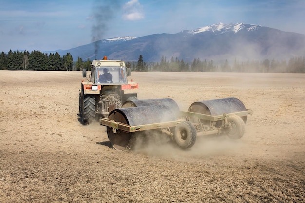 Traktor zieht Schwermetallwalze auf trockenem Feld, Staub dahinter und schwarzer Rauch oben, Berge im Rücken. Frühling Bodenvorbereitung.