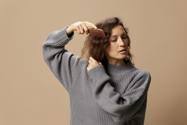 Foto tranquila, bastante rizada, hermosa mujer con suéter casual gris con cepillo para el cabello, peines, el cabello se ve a un lado posando aislado sobre un fondo pastel beige problemático concepto de cabello dañado rebelde copiar espacio