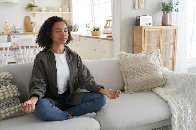 Tranquila chica de raza mixta hace yoga en casa sentada en posición de loto en el sofá Meditación estilo de vida saludable