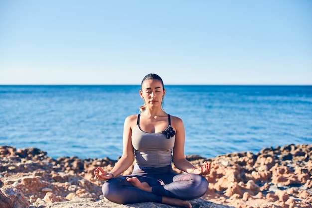 Tranquila joven meditando en la playa contra un hermoso mar azul.