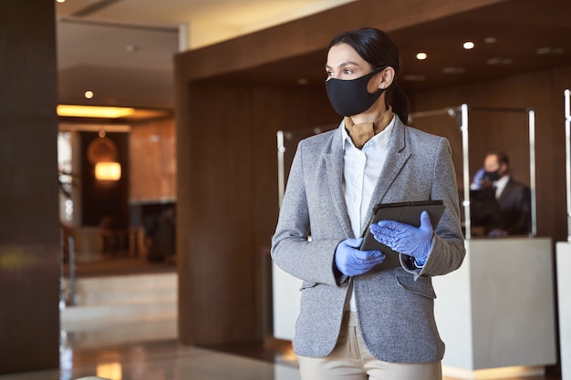 Tranquila joven de pie con una tableta moderna en el vestíbulo de un hotel vestida de acuerdo con las precauciones de seguridad