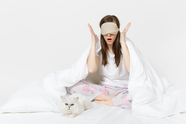 Foto tranquila joven sentada en la cama con antifaz para dormir en los ojos aislados en la pared blanca