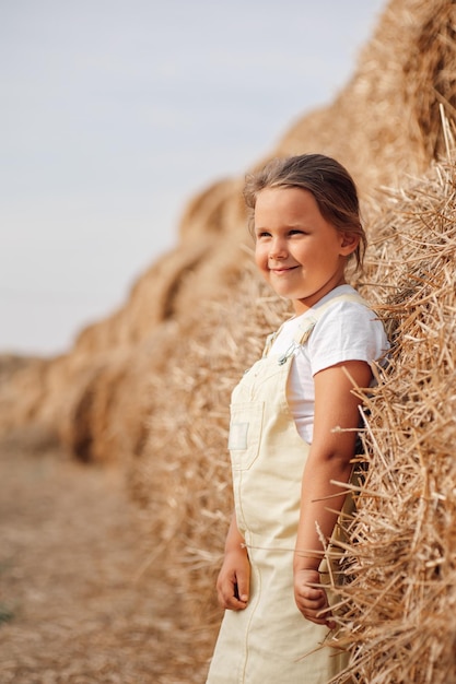 Tranquila, linda, sonriente, feliz niña mirando hacia otro lado apoyándose en un enorme pajar en el campo, manos tocando heno con los ojos entrecerrados Tiempo lejos de la ciudad en el campo rural