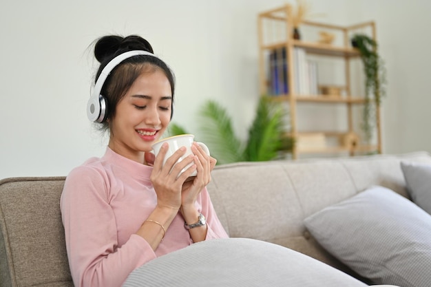 Tranquila mujer asiática escuchando música mientras huele su café y se relaja en el sofá