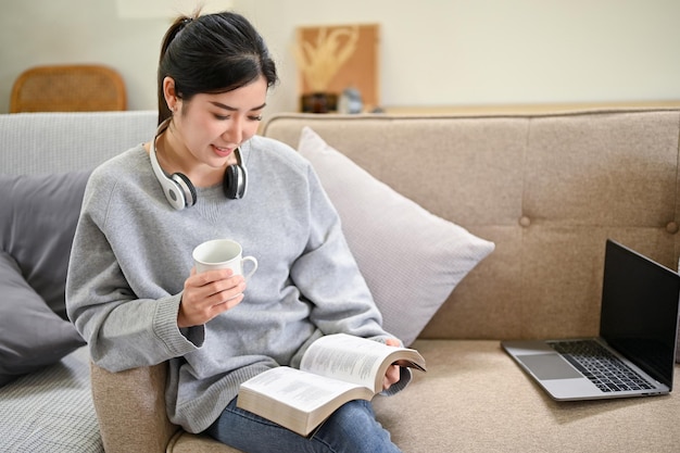 Tranquila mujer asiática milenaria leyendo un libro y bebiendo café mientras se relaja en la sala de estar