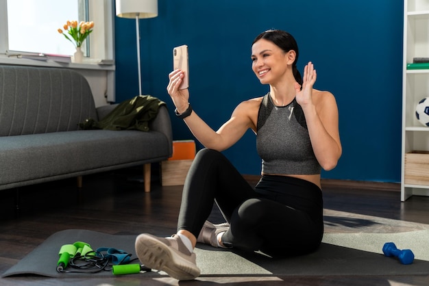 Tranquila mujer flexible en ropa deportiva sentada en la estera en posición de loto y haciendo yoga mientras tiene video chat en el teléfono inteligente en casa