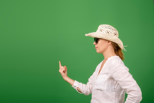 Tranquila mujer de mediana edad segura de sí misma con una gorra, camisa blanca y gafas de sol sobre un fondo verde. Se pone de lado y señala con el dedo el lugar del texto.