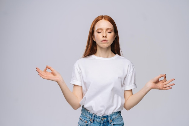 Tranquila y serena mujer joven con camiseta y pantalones de mezclilla manteniendo los ojos cerrados mientras medita, sintiéndose relajada, tranquila y pacífica, tomándose de la mano con un signo de mudra en un fondo blanco aislado.