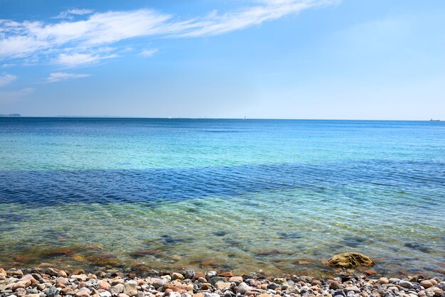 Las tranquilas olas del océano se lavan sobre las piedras en una playa vacía con veleros navegando en el horizonte Paisaje escénico para unas relajantes vacaciones de verano Copie el espacio en el mar con un fondo de cielo azul