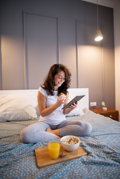 Tranquilo, bastante activo de mediana edad disfrutando de la cena y el jugo mientras lee noticias de una tableta en la cama.