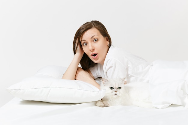 Tranquilo joven acostado en la cama con blanco lindo gato chinchilla plateado persa, sábana, almohada, manta en la pared blanca